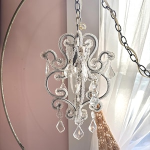 May include: A small, ornate chandelier with clear crystal accents and teardrop-shaped pendants. The chandelier has a silver-colored metal frame and is suspended by a chain. The design is vintage and elegant, suitable for home decor.