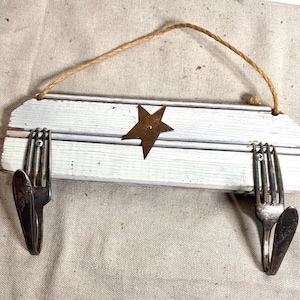 May include: A rectangular white-painted wooden wall hook rack featuring a centered brown metal star and two silver-toned metal forks bent into hooks, attached with screws. A thin brown twine cord is looped through the top for hanging.