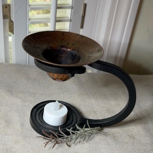 May include: A black metal oil warmer featuring a curved arm and a circular base holding a white tea light candle. A hammered copper bowl sits at the top of the arm. A small sprig of dried greenery rests on the base near the candle.
