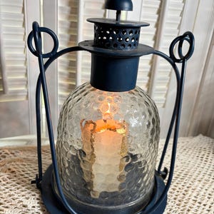 May include: A dark blue metal lantern featuring a clear, textured glass globe with a dimpled pattern. The lantern has a circular base, a perforated top chimney, and a curved handle. Inside the glass globe, a single lit candle glows warmly.