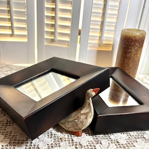 May include: Two square mirrors with dark brown, matte-finished frames are positioned at an angle. A small, hand-painted wooden goose figurine with a light brown body and a bright orange beak stands between the two mirror frames.