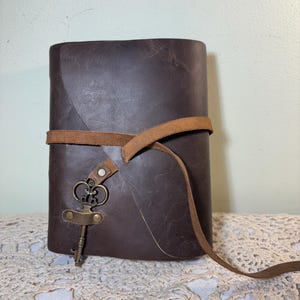 May include: A dark brown leather journal with a wrap-around strap closure. The journal features a decorative antique-style key charm. The journal is sitting on a cream-colored lace tablecloth.