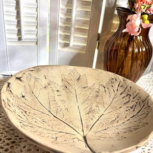 May include: A shallow, round, beige ceramic bowl featuring a dark brown, detailed leaf imprint across its interior surface. The bowl has an organic, slightly uneven rim and a matte texture, showcasing the intricate veins of the leaf design.