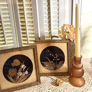 May include: Two square gold-toned frames containing circular pressed flower arrangements on dark backgrounds, accompanied by a single dried rose and a tall, frosted amber-colored glass candle holder with a long, thin cream-colored taper candle.