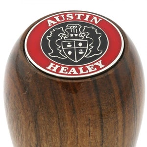 May include: A wooden gear knob with a red and black emblem that reads "AUSTIN HEALEY". The knob is made of dark brown wood with visible grain. The emblem features a crest design.