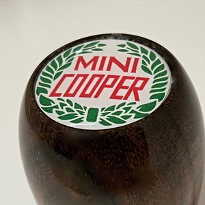 May include: A brown wooden gear shift knob with a white circular top featuring a green laurel wreath and the text "MINI COOPER" in red letters.