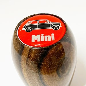 May include: A wooden gear stick knob with a glossy finish. The top has a red circle with a black car graphic and the word "Mini" in white. The knob has a dark brown wood grain pattern.