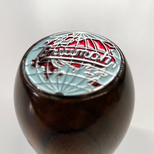 Op de afbeelding: Een houten versnellingspookknop met een rood en zilveren globe-embleem met het woord "Triumph" erop.