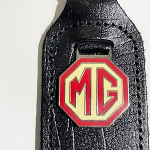 Op de afbeelding: Zwarte leren sleutelhanger met een zilveren metalen MG-logo. Het logo is rood en geel met een witte omtrek.
