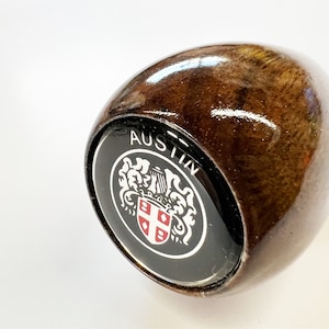 Puede incluir: Pomo de cambio de madera con acabado brillante en color marrón oscuro. El pomo presenta un emblema negro con la palabra "AUSTIN" y un diseño de escudo con detalles rojos y blancos.