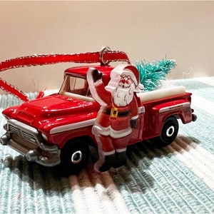 Puede incluir: Adorno de Navidad de camión rojo con una figura de Papá Noel sosteniendo un pequeño árbol de Navidad verde. El camión tiene una franja blanca y las palabras "Matthews" y "10" en el lateral.