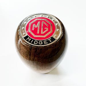 Puede incluir: Una palanca de cambios de madera con un logotipo MG Midget plateado y rojo.