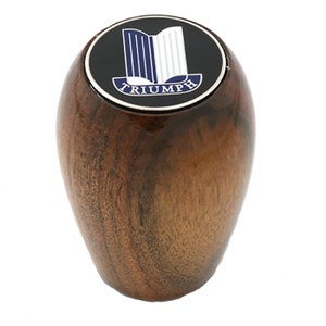 May include: A wooden gear shift knob with a black and silver Triumph logo on top. The logo features a blue and white stylized design.