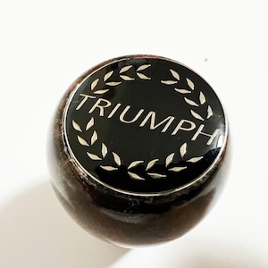 Può includere: Oggetto rotondo marrone scuro con una parte superiore nera. La parte superiore presenta la parola "TRIUMPH" in bianco, circondata da un design a corona d'alloro. L'oggetto ha una finitura lucida ed è posto su uno sfondo bianco.