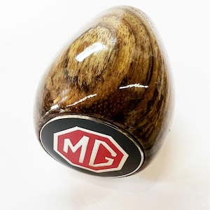 May include: A wooden gear shift knob with a black and red MG logo. The knob is brown with a wood grain pattern.