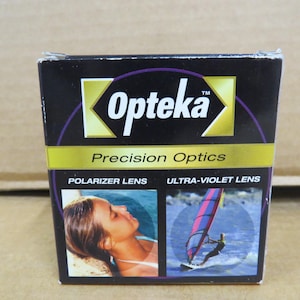 Può includere: Scatola nera con il marchio "Opteka" in lettere bianche. La scatola presenta le parole "Precision Optics", "Polarizer Lens" e "Ultra-Violet Lens". Sulla scatola sono presenti anche immagini di una persona e di un windsurfista.