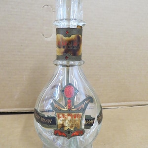 May include: Clear glass liquor bottle with a handle and a decorative label. The label features text and a crest design. The bottle has a bulbous body and a cylindrical neck. The label reads "Apricot Brandy".