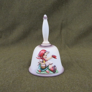 May include: A white ceramic bell with a red and white illustration of a girl in a red hooded dress holding a red bird. The girl is sitting on a green patch of grass with red flowers.