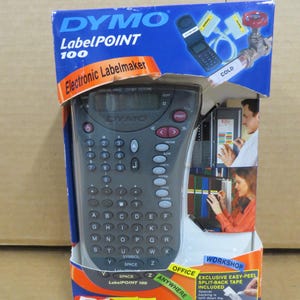 Puede incluir: Una rotuladora electrónica Dymo LabelPOINT 100 en su embalaje original. La rotuladora es gris con un teclado QWERTY y una pequeña pantalla. El embalaje es azul y blanco con información e imágenes del producto.