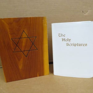 May include: Two books are displayed. One is a wooden book with a Star of David etched into the front. The other is a white book with the words "The Holy Scriptures" printed on the front.