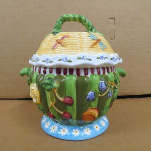 May include: A decorative ceramic jar with a lid and handle. The jar features a green and white striped body, a yellow woven-style lid with dragonfly accents, and a blue base with white daisy details. The jar is adorned with floral and fruit motifs.