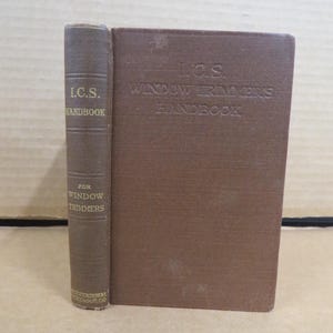 May include: A vintage brown book titled "I.C.S. Handbook for Window Trimmers." The book has a textured cover and spine, with the title embossed in a darker shade. The book is standing upright.
