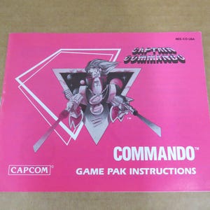 Puede incluir: Manual de instrucciones rosa del juego Captain Commando. La portada presenta una ilustración de personaje dentro de un diseño geométrico blanco. El texto "CAPTAIN COMMANDO" y "COMMANDO" se muestran, junto con el logotipo de Capcom.