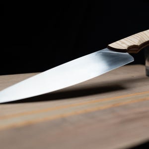 May include: A large, stainless steel chef's knife with a light-colored wooden handle rests on a wooden cutting board. A small glass is in the background. The knife blade is angled, and the background is black.