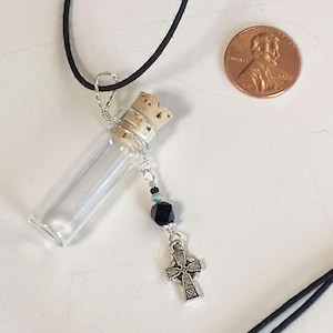 May include: A clear glass vial necklace with a cork stopper, a silver cross charm, and black cord. The cross charm has an intricate design. A penny is next to the necklace for size reference.