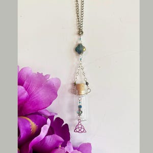 Può includere: Un flacone di vetro trasparente con un tappo di sughero, sospeso da una catena d'argento e ornato di perline. Un ciondolo con nodo celtico d'argento è appeso sotto. Il flacone è lungo circa 5 cm.