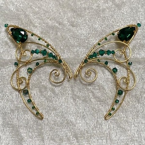 May include: A pair of gold-toned wire elf ear cuffs. Each cuff features a large, teardrop-shaped, emerald-green gemstone and smaller green beads. The wire is intricately shaped with swirls and curves, creating a delicate, fantasy-inspired accessory.