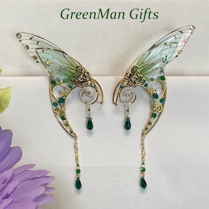 May include: A pair of handcrafted fairy ear cuffs with delicate, translucent green wings and intricate gold-toned wirework. The design features sparkling green and clear crystals, with dangling emerald green teardrop beads. The text "GreenMan Gifts" is at the top.