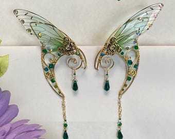 May include: A pair of handcrafted fairy ear cuffs with delicate, translucent green wings and intricate gold-toned wirework. The design features sparkling green and clear crystals, with dangling emerald green teardrop beads. The text "GreenMan Gifts" is at the top.