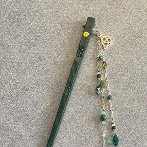 Pode incluir: Um palito de cabelo de madeira verde escuro com detalhes decorativos. O palito apresenta um pequeno adorno dourado e é adornado com uma corrente pendurada de contas verdes e um pingente de nó celta prateado. Acessório de cabelo elegante.