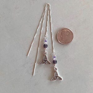 May include: A pair of silver earrings with a purple bead and a silver Celtic knot charm. The earrings are hanging from a silver chain.