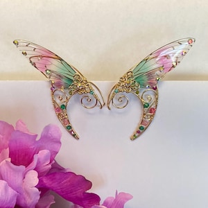 May include: Gold-coloured fairy ear cuffs with translucent wings in pink and green. The wings are embellished with colourful gemstones. The ear cuffs have intricate filigree detailing, creating a fantasy aesthetic.
