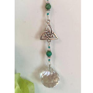 May include: A silver Celtic knot charm hangs from a chain with green and clear beads. A clear crystal hangs from the bottom of the charm.