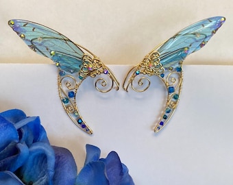 May include: A pair of gold-toned elf ear cuffs with iridescent blue butterfly wing accents. The cuffs are adorned with blue and iridescent crystals. The design features intricate wirework and decorative filigree.