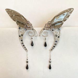 May include: A pair of silver-toned elf ear cuffs with iridescent wing-shaped accents. The cuffs feature black bead and teardrop dangles, with decorative filigree and clear crystal accents. These fantasy-themed ear cuffs are designed to be worn on the ears.