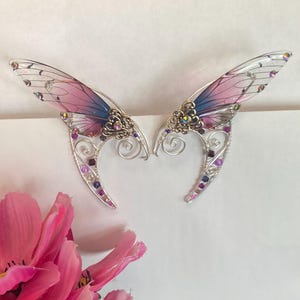 May include: A pair of ear cuffs featuring translucent butterfly-style wings with a gradient of pink and blue. The silver-toned wire frame is decorated with intricate metal filigree, swirling wire accents, and small purple and clear crystals.