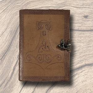 May include: A brown leather journal with a decorative border and a metal clasp. The cover features an embossed design of Thor's hammer, Mjolnir, and Celtic knotwork. The journal is closed and sits on a wooden surface.