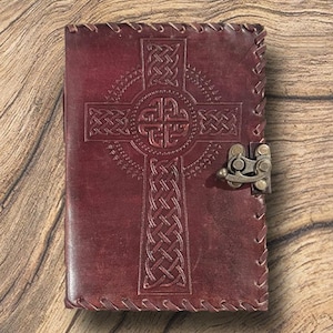 May include: A burgundy leather journal with a Celtic cross design embossed on the cover. The journal features a metal clasp closure and stitched edges. The cover is a deep red-brown color.