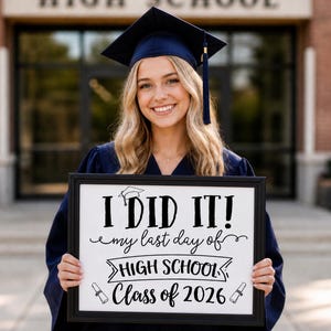 Puede incluir: Una graduada sonriente con birrete y toga azul marino sostiene un letrero enmarcado. El letrero dice "I DID IT! my last day of HIGH SCHOOL Class of 2026" en letras negras. El fondo muestra un edificio de ladrillo.