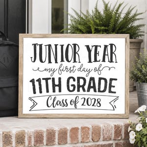 May include: A rectangular sign with a light wood frame featuring black text on a white background that reads: JUNIOR YEAR my first day of 11TH GRADE Class of 2028. The sign is positioned next to a black backpack and green potted plants.