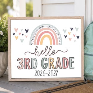 May include: A rectangular sign in a light wood frame featuring a rainbow graphic, small hearts, and the text "hello 3RD GRADE 2026-2027" in a mix of muted pink, blue, and yellow colors. The sign is displayed next to a light green backpack.
