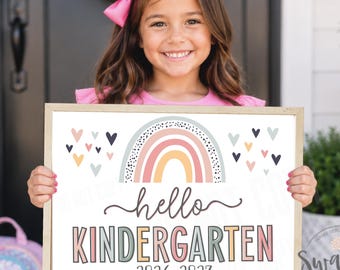 May include: A rectangular sign with a light wood frame featuring a colorful rainbow illustration, small heart icons, and the text "hello KINDERGARTEN 2026-2027" in a mix of script and block lettering. The design uses a palette of muted pink, yellow, and blue.