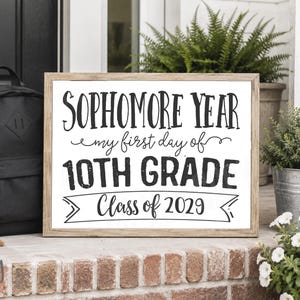 May include: A rectangular sign with a light wood frame featuring black text on a white background that reads: SOPHOMORE YEAR my first day of 10TH GRADE Class of 2029. The sign is positioned next to a black backpack and green potted plants.