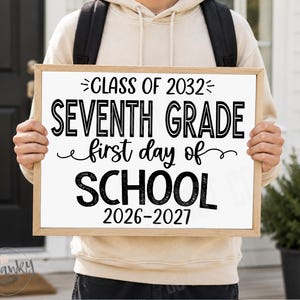 May include: A rectangular sign with a light wood frame featuring black text on a white background that reads: CLASS OF 2032, SEVENTH GRADE, first day of, SCHOOL, 2026-2027. The sign is held up, displaying the school-themed announcement clearly.