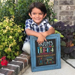 May include: A young child smiles while holding a blue framed chalkboard. The chalkboard reads "Jatum's First Day of Pre-K 4" and includes the date, school name, and teacher's name. The child is wearing a blue and white checkered shirt.