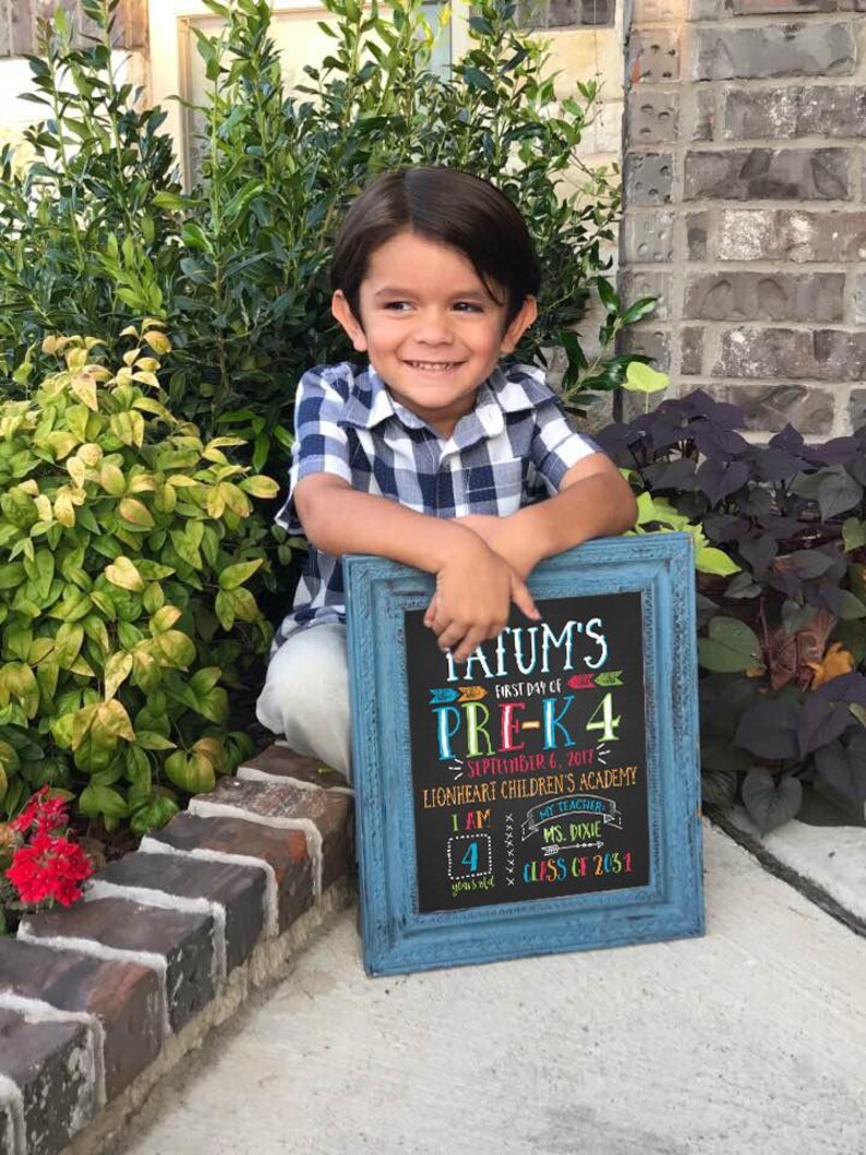 May include: A young child smiles while holding a blue framed chalkboard. The chalkboard reads "Jatum's First Day of Pre-K 4" and includes the date, school name, and teacher's name. The child is wearing a blue and white checkered shirt.